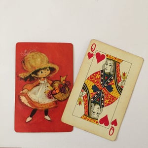 May include: Two playing cards. One card features a vintage illustration of a girl in a sun hat carrying a basket of fruit. The other card is the Queen of Hearts from a standard deck of cards.