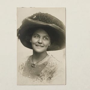 May include: A black and white vintage photograph of a woman wearing a large feathered hat and a patterned dress. She is smiling and looking to the right of the frame. The photo is signed "C. Helen Brown, Chicago."
