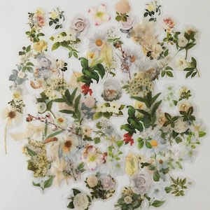 以下が含まれることがあります： 緑の葉が付いた白とクリーム色の花のステッカーのコレクション。ステッカーはクラスター状に配置されており、バラ、デイジー、ユリなど、さまざまな種類の花が特徴です。