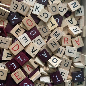 May include: A collection of square wooden game tiles featuring black and red letters with numerical values printed on each piece. The tiles are scattered in a jumbled pile, displaying various characters including D, N, K, O, W, A, R, S, T, and Y.