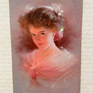 May include: Vintage postcard featuring a portrait of a woman with a soft, romantic aesthetic. The woman has brown hair, adorned with flowers, and wears a pink dress. The postcard has a faded, aged appearance.