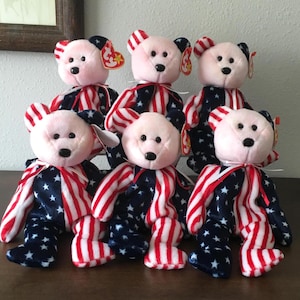 May include: Six plush teddy bears with pink faces and ears, black button eyes, and black noses. The bears wear outfits with a blue star pattern and red and white striped arms and legs. Each bear has a red ribbon around its neck.