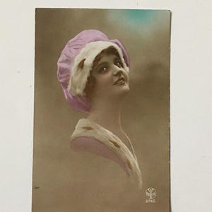 May include: A vintage postcard featuring a woman wearing a light purple and white hat. The woman is looking up and smiling. The postcard has a brown background and a blue and green sky in the background. The postcard is marked with the number 2416.