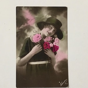May include: A vintage postcard featuring a woman in a black hat and dress holding a bouquet of pink and red roses. The background is a soft, faded pink and brown.