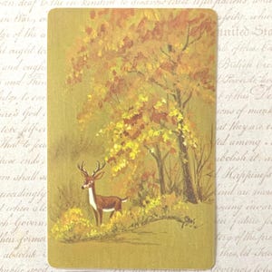 May include: A rectangular playing card featuring a painted illustration of a brown deer with antlers standing in a forest setting with yellow and orange autumn foliage on the trees and ground, set against a muted gold-toned background.