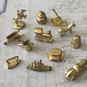 May include: A set of 11 gold-colored metal game pieces, including a thimble, a boot, a wheelbarrow, a ship, a hat, a train, a dog, a  person on a horse, a  cannon, an iron, and a  bag. The pieces are arranged on a white surface with handwritten text.