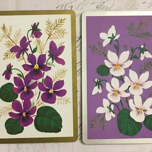 May include: Two vintage playing cards with a floral design. The card on the left has purple flowers on a white background with gold trim. The card on the right has white flowers on a purple background with silver trim.