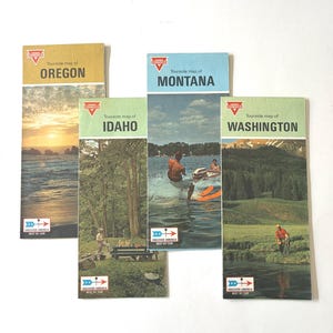 May include: Four vintage Conoco Touraide travel maps for Oregon, Idaho, Montana, and Washington. Each rectangular paper map features a color photograph of a regional landscape or activity, the state name, and the Discover America Best By Car logo.