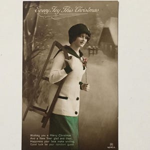 May include: Vintage Christmas postcard featuring a woman in a white coat and green skirt holding a wooden sled. The text on the postcard reads "Every Joy This Christmas" and includes a holiday greeting.