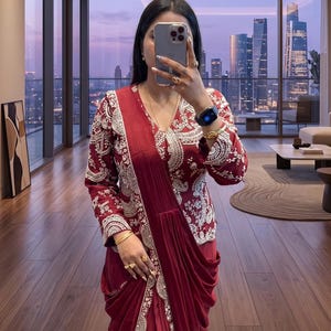 May include: A deep maroon ensemble featuring a long-sleeved jacket with intricate white embroidery and a matching draped skirt. The jacket has a V-neckline and detailed patterns along the cuffs and hem, while the skirt features soft folds.