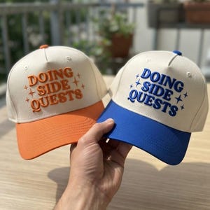 May include: Two baseball caps with off-white crowns and colored brims. One cap has an orange brim and orange embroidered text reading DOING SIDE QUESTS with star accents. The other cap has a blue brim and blue embroidered text reading DOING SIDE QUESTS.