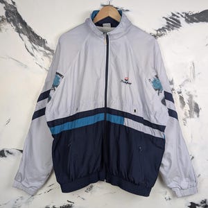 Puede incluir: Una chaqueta cortavientos de color gris claro con cremallera completa de color azul oscuro, cuello alto y mangas largas. La chaqueta presenta una franja horizontal azul oscuro y azul verdoso en el pecho, con detalles estampados en los hombros.