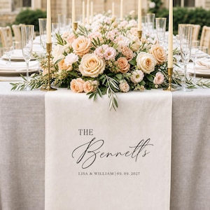May include: A cream-colored fabric table runner featuring the black printed text THE Bennetts LISA & WILLIAM | 09. 09. 2027. The runner is displayed on a table set with a floral centerpiece of peach and pink roses, tall taper candles, and clear glasses.