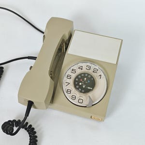 Könnte beinhalten: Ein beiges Wählscheibentelefon mit passendem Hörer und einem schwarzen Spiralkabel. Das Gerät verfügt über eine kreisförmige Wählscheibe mit schwarzen Ziffern von null bis neun und ein leeres weißes rechteckiges Etikettenfeld.