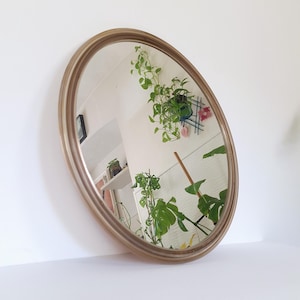 Puede incluir: Un espejo redondo con un marco liso de color dorado metálico. La superficie reflectante muestra una vista clara del interior de una habitación, incluyendo plantas verdes en macetas, una pared blanca y un pequeño cuadro en el fondo.