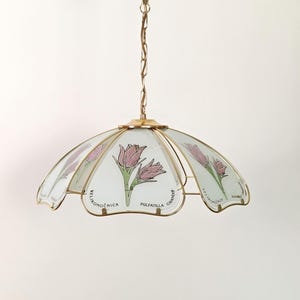 Könnte beinhalten: Eine Hängelampe mit einem goldfarbenen Metallrahmen und einer Kette. Der Schirm besteht aus gebogenen Glasscheiben mit Illustrationen von rosa Blumen mit grünen Stielen. Der Text "VELIKONOČNICA" und "PULSATILLA GRANDIS" ist auf dem Glas aufgedruckt.