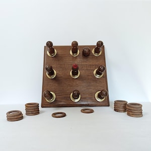 Pode incluir: Um jogo de tabuleiro quadrado de madeira com uma grade de nove pinos verticais com bases na cor latão. Um pino tem o topo vermelho e uma pequena bola de madeira repousa sobre o tabuleiro. Vários anéis de madeira estão empilhados e espalhados.