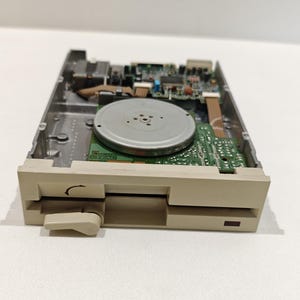 Puede incluir: Una disquetera TEAC de color beige con una placa de circuito interno expuesta, un componente de motor metálico circular y una palanca de expulsión frontal. El dispositivo cuenta con una ranura horizontal y una pequeña luz indicadora rectangular.