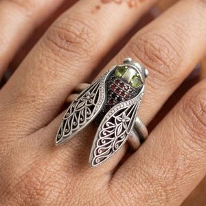 Può includere: Un anello in argento a forma di cicala, caratterizzato da intricati motivi a filigrana sulle ali, una testa incastonata con due piccole pietre verdi e un torace decorato con numerose minuscole pietre rosso scuro su una fascia lucida.