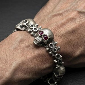 Könnte beinhalten: Ein silberfarbenes Metallarmband mit einer Reihe detaillierter Totenkopfmotive, die durch Unendlichkeitssymbole verbunden sind. Der mittlere Totenkopf ist mit zwei kleinen, runden, dunkelroten Steinen in den Augenhöhlen akzentuiert.