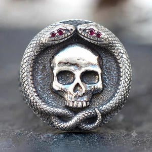 May include: A circular silver ring featuring a central human skull motif surrounded by two intertwined snakes. The snakes have detailed scales and small, vibrant red gemstone eyes. The metal has a dark, oxidised finish that highlights the texture.