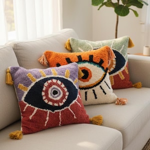 May include: Three square throw pillows featuring tufted eye designs with colorful patterns. Each pillow has yellow tassels at the corners. The front pillow is purple, red, and dark blue. The middle pillow is orange and white. The back pillow is green.