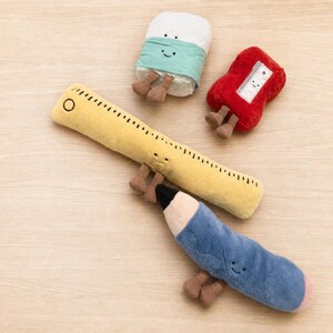 Puede incluir: Un conjunto de cuatro peluches con forma de material escolar, que incluye una regla amarilla, un lápiz azul, una goma de borrar blanca con una banda turquesa y un sacapuntas rojo. Cada juguete tiene una carita bordada y pies marrones.