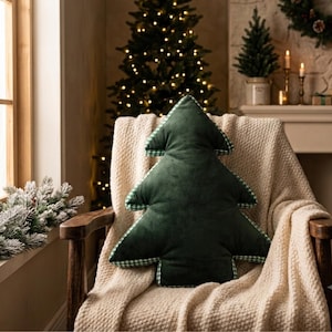 Könnte beinhalten: Ein dunkelgrünes Samtkissen in Form eines Weihnachtsbaums mit einem weiß-grün karierten Stoffbesatz an den Rändern. Das Kissen liegt auf einer strukturierten cremefarbenen Decke, die über einen Holzstuhl drapiert ist.