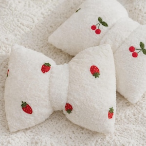 Peut inclure: Deux nœuds pour cheveux en peluche blanche avec une texture douce et duveteuse. Chaque nœud est décoré de fraises rouges brodées avec des feuilles vertes, ainsi que de petites cerises rouges avec des tiges vertes sur le tissu.