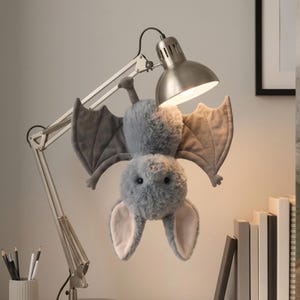 Op de afbeelding: Een pluche vleermuis met zachte grijze vacht, grote lichtroze binnenoren en donkergrijze vleugels, ondersteboven hangend aan de metalen arm van een zilveren bureaulamp. De vleermuis heeft kleine zwarte oogjes en een roze neusje.