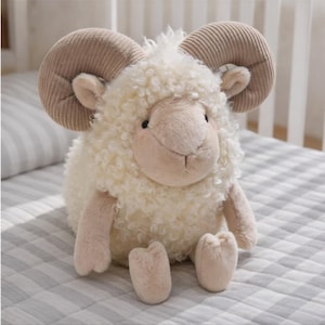 May include: A plush stuffed animal in the shape of a ram with cream-colored curly faux fur, light tan corduroy-textured curved horns, and matching tan fabric on the face, ears, arms, and feet, featuring small black eyes and a stitched nose.