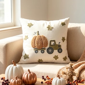 May include: A square off-white throw pillow featuring a textured, raised embroidery design of a tan pumpkin sitting on a trailer pulled by an olive green tractor, surrounded by small embroidered olive green leaves scattered across the fabric.