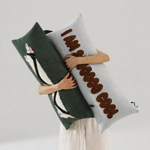 May include: Two rectangular throw pillows. One is dark green with a white and black embroidered animal graphic. The other is light grey with brown tufted text that reads "I'M SOOOOO COOL" in a vertical orientation, featuring small black side tabs.