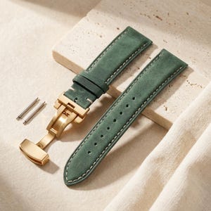 Puede incluir: Una correa de reloj de cuero verde bosque de dos piezas con un cierre desplegable de metal dorado y costuras blancas en contraste. El conjunto incluye dos pequeños pasadores de resorte de metal, con una textura mate y un diseño cónico.