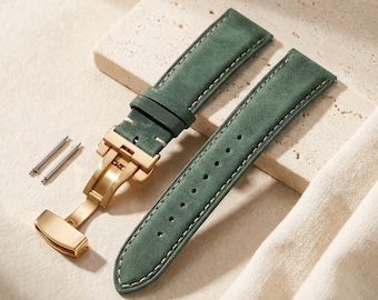 May include: A two-piece forest green leather watch strap featuring a gold-toned metal deployment clasp and white contrast stitching. The set includes two small metal spring bars for attachment, showcasing a matte texture and a tapered design.