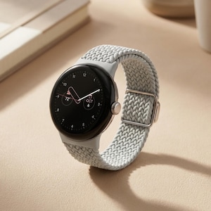 May include: A smartwatch with a circular black display showing the time, date, and temperature, paired with a light gray braided fabric band. The watch features a metallic side crown and a silver-toned rectangular buckle on the woven strap.