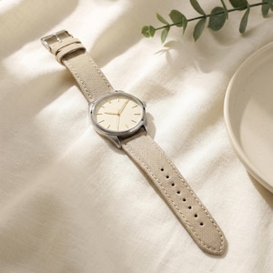 May include: A wristwatch featuring a round silver-tone metal case and a cream-colored dial with simple hour markers. The watch is attached to a textured beige canvas strap with a silver-tone metal buckle and multiple adjustment holes.