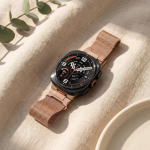 May include: A smartwatch with a black rectangular case and a rose gold-toned metal mesh band. The digital watch face displays a complex interface with white numerals, orange accents, and various data indicators including a battery level of 27 percent.