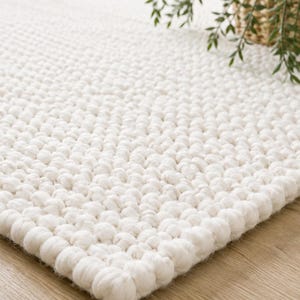 May include: A rectangular area rug featuring a thick, chunky knit texture in a solid off-white color. The rug is constructed from individual soft, rounded yarn loops arranged in a dense, uniform pattern across the entire surface of the piece.