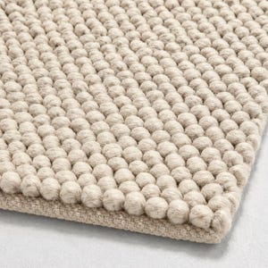 Può includere: Un tappeto beige caratterizzato da una trama densa e uniforme composta da numerose piccole sfere di lana infeltrita arrotondate, disposte in una griglia stretta, con un bordo in tessuto piatto visibile lungo il lato inferiore del pezzo.