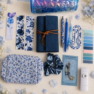 Könnte beinhalten: Eine Sammlung blauer Schreibwaren, darunter ein facettierter, irisierender Becher, ein dunkelblaues Notizbuch mit Lederumschlag und Blattanhänger, florale Lesezeichen, drei Stifte, ein blaues Blumenetui, ein Satin-Haargummi und Klebestreifen.