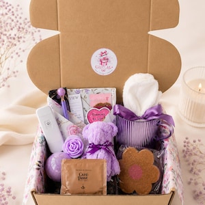 Könnte beinhalten: Eine Geschenkschachtel aus Pappe mit einer gerippten lila Glastasse mit weißem Handtuch, einem kleinen lila Plüschbären, einem blumenförmigen Keks, einer Café-Tasse Schokoladenpackung, einem lila Stift, einem weißen elektronischen Gerät und einer lila Badebombe.
