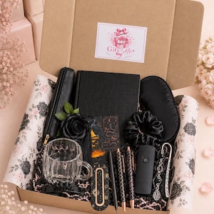 Könnte beinhalten: Eine Geschenkbox mit einem schwarzen Notizbuch mit strukturierter Oberfläche, einer klaren Glastasse, einer schwarzen Schlafmaske, einem schwarzen Haargummi, zwei Stiften, einer schwarzen Rose, zwei Lesezeichen, einem schwarzen Gerät und einem Metall-Lesezeichen.