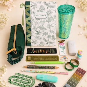 Può includere: Un astuccio verde, un taccuino con illustrazioni di fiori e uccelli, un bicchiere testurizzato turchese con cannuccia, due segnalibri, un telecomando bianco, una gomma arcobaleno, due rotoli di nastro adesivo verde, penne, una matita, una grande graffetta verde, un evidenziatore, un set di linguette adesive e un elastico per capelli nero.