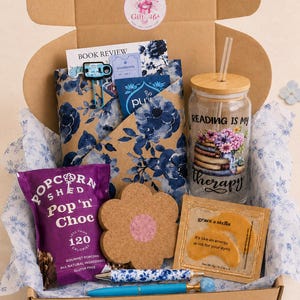 Op de afbeelding: Een kartonnen geschenkdoos met een glazen beker, bamboe deksel en rietje, bedrukt met boeken, bloemen en de tekst Reading is my therapy. Bevat een paarse zak Pop 'n' Choc popcorn, een bloemvormige kurken onderzetter, twee pennen en oogmaskers.