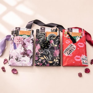 Può includere: Tre pacchetti regalo rettangolari avvolti in carta fantasia. Quello a sinistra è viola chiaro con fiori, quello centrale è nero con fiori e quello a destra è rosso con labbra e il testo THRILLER GIRLY e I love THRILLER Books.