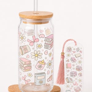Può includere: Un bicchiere in vetro trasparente con coperchio in bambù e cannuccia in vetro, decorato con un motivo di libri rosa, fiori, fiocchi e candele. Un segnalibro rettangolare abbinato con una nappa rosa e illustrazioni identiche è posto accanto.