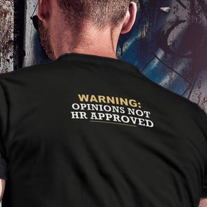 May include: A black short-sleeved t-shirt featuring the text WARNING: OPINIONS NOT HR APPROVED printed in white and gold on the back. The shirt has a crew neckline and a plain black fabric construction, displaying the text clearly across the upper back.