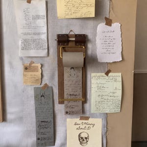 May include: A wooden wall-mounted paper roll holder with a brass bar and brown leather straps, holding a roll of paper with handwritten text. The holder is surrounded by various vintage-style documents, notes, and a skull illustration on paper.