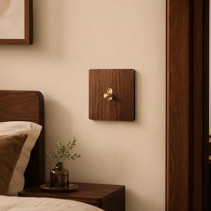 May include: A square light switch cover plate made of dark brown wood with a prominent vertical grain pattern. Centered on the plate is a small, polished gold-toned metal toggle switch with a rounded, elongated base plate.